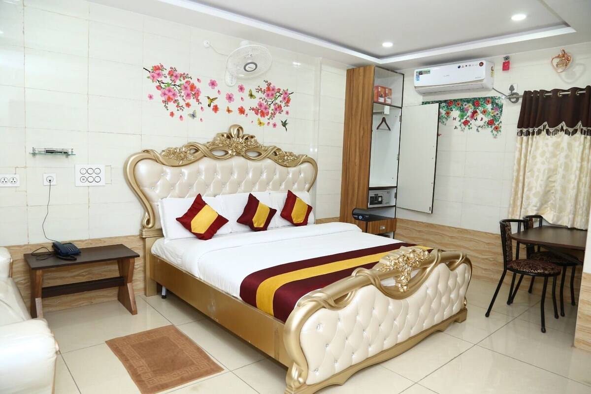 A hotel room with a large bed, a bedside table, a desk, a chair, an air conditioning unit, and a wall-mounted fan.