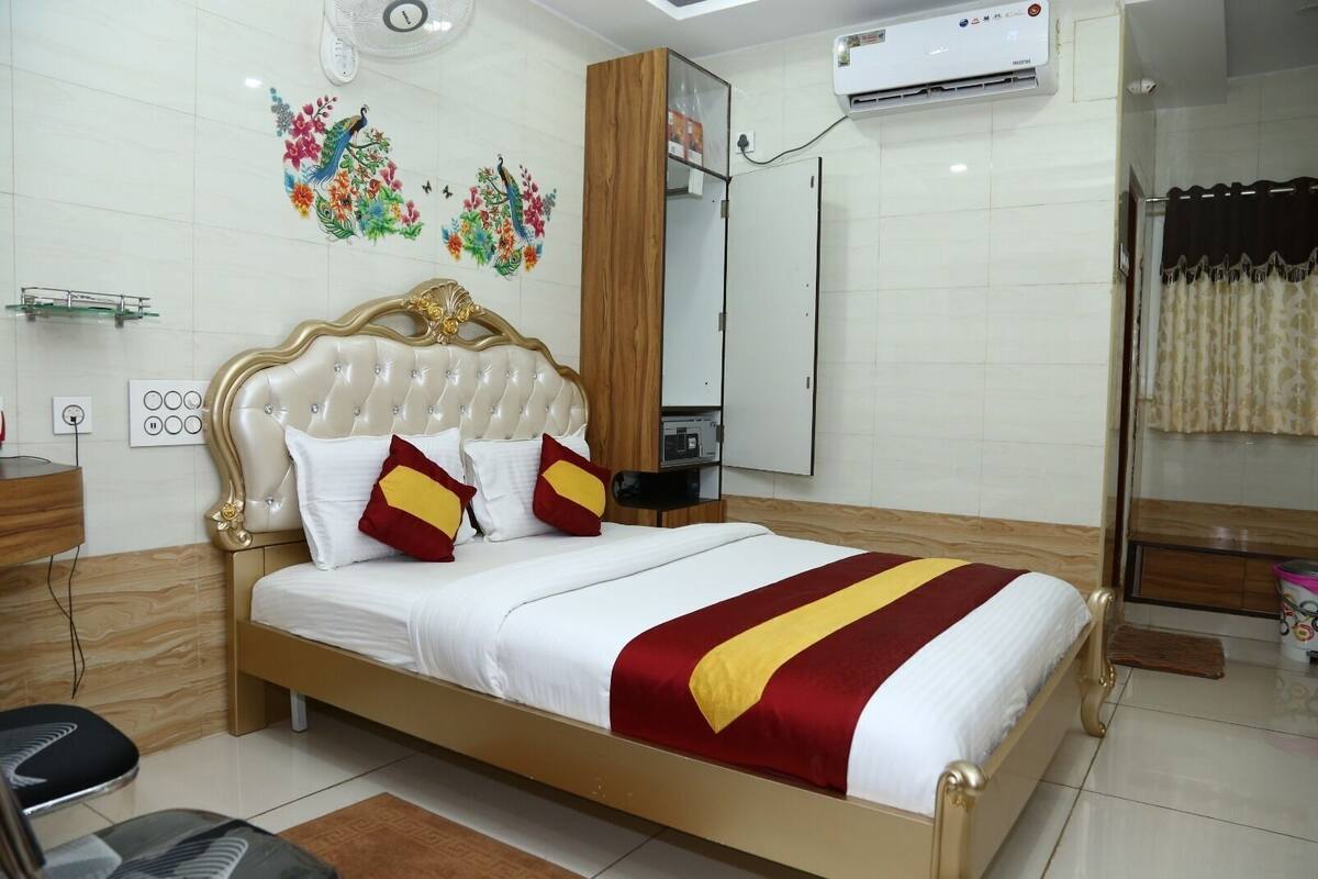 A neatly arranged hotel room with a bed, a bedside table, a mirror, a wall-mounted air conditioner, and a decorative floral wall art piece.