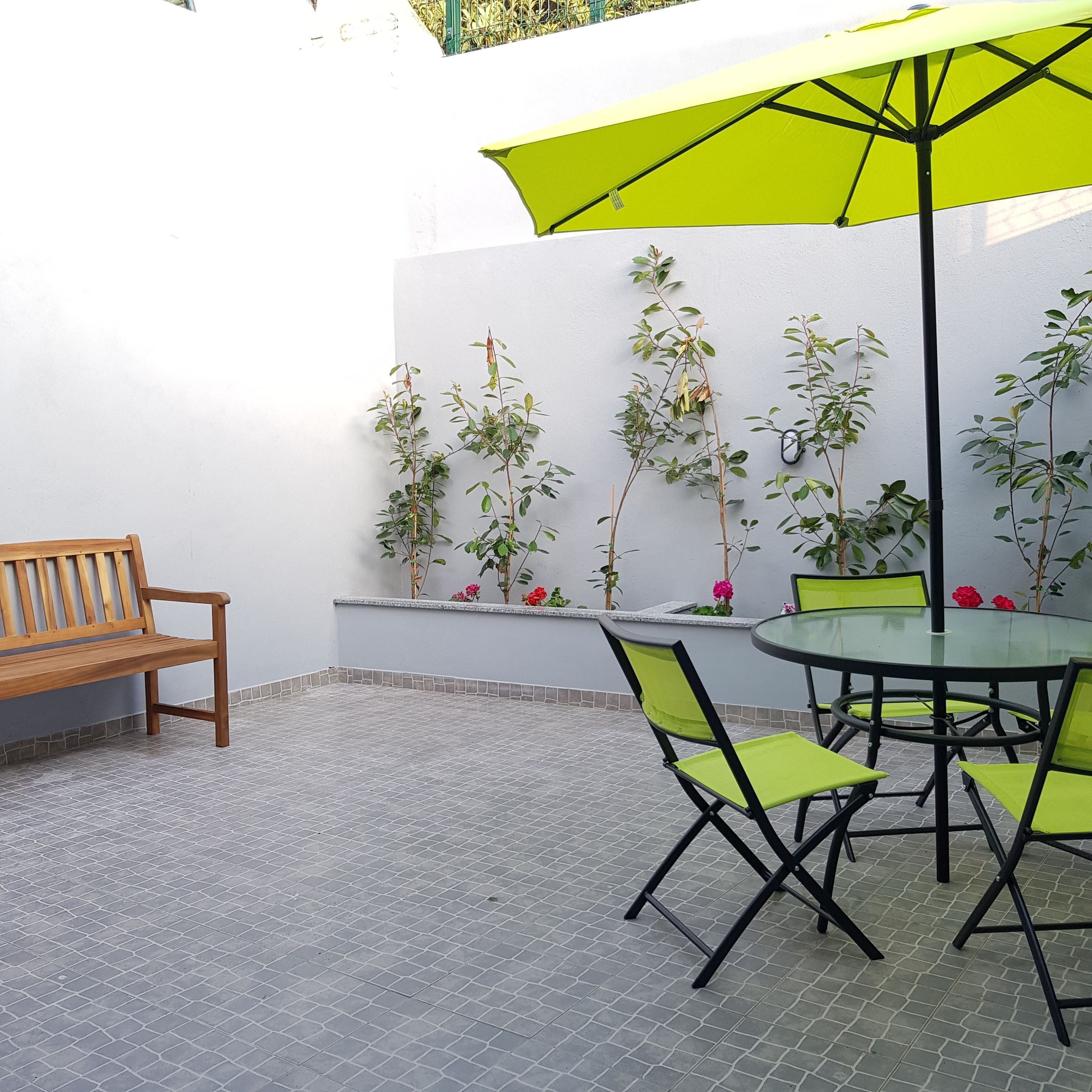 Un patio con una mesa redonda de vidrio, sillas verdes, un banco de madera y un gran paraguas verde.