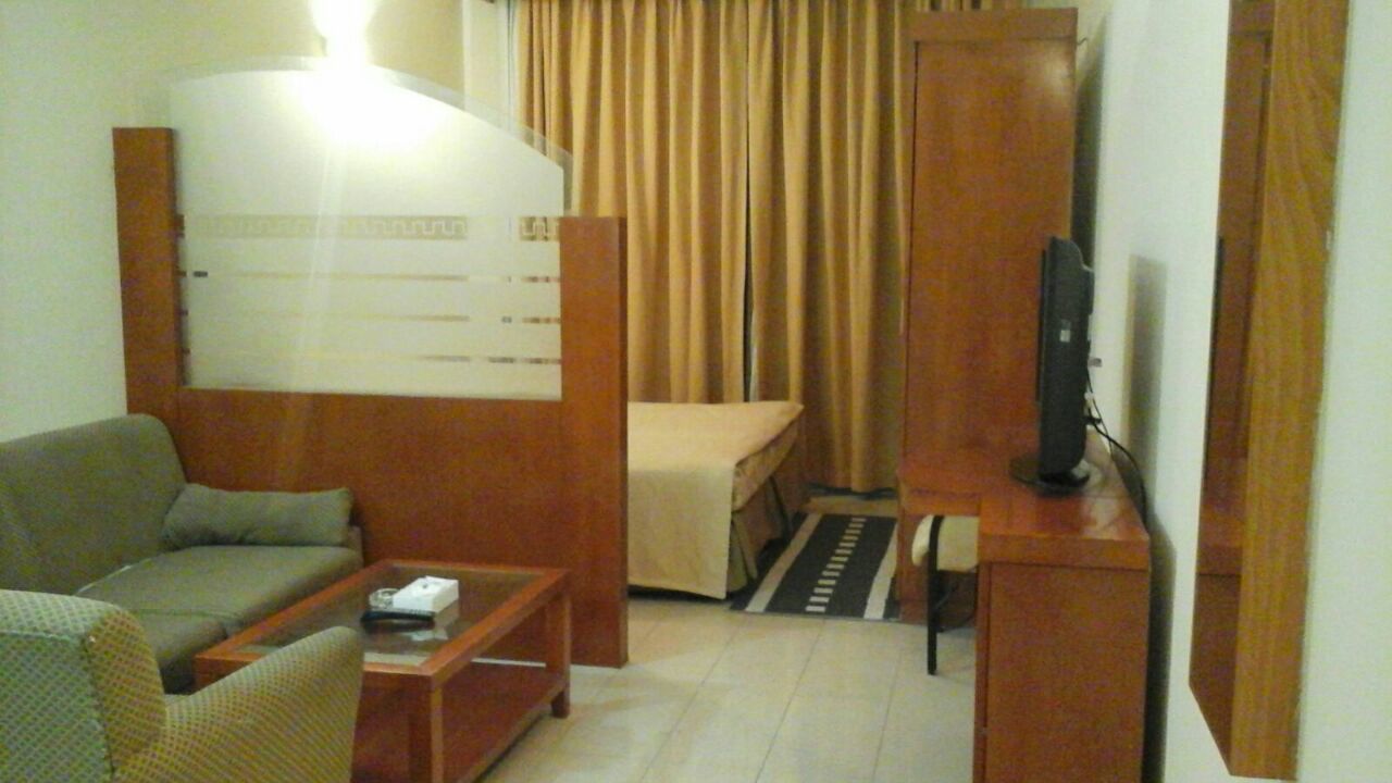 A compact hotel room with a sofa, a small table, a television, and a bed with curtains.