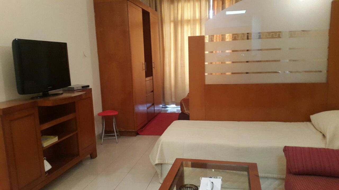 A hotel room with a bed, a television on a wooden stand, a small table with a glass top, and a red stool.