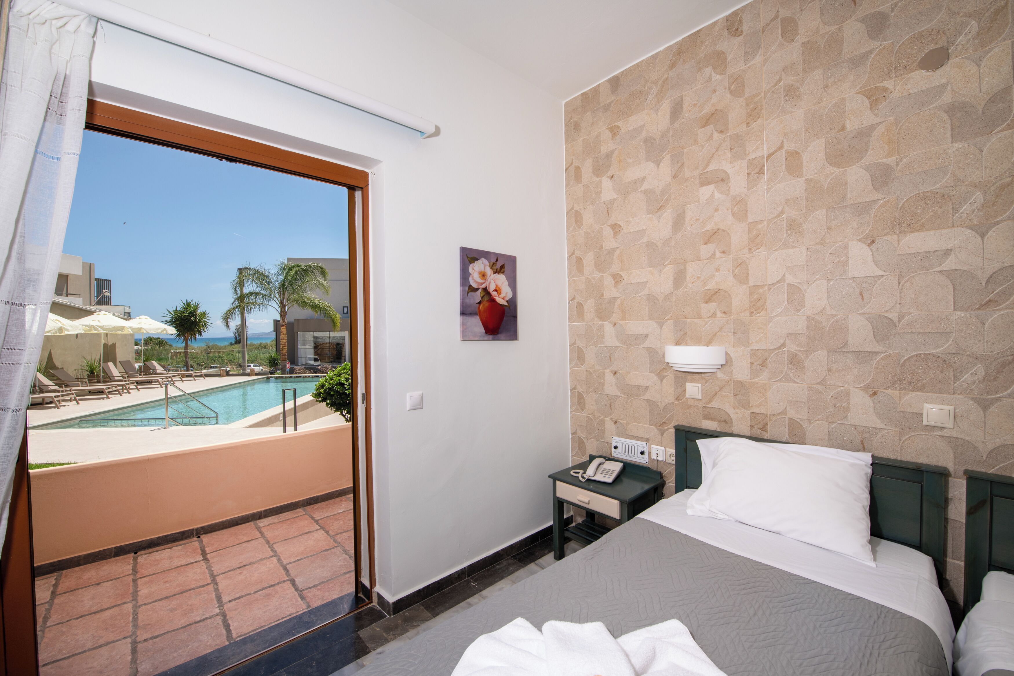 A hotel room with a bed, a nightstand, a telephone, a painting, and a view of a swimming pool and palm trees.