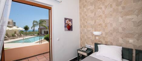 A hotel room with a bed, a nightstand, a telephone, a painting, and a view of a swimming pool and palm trees.