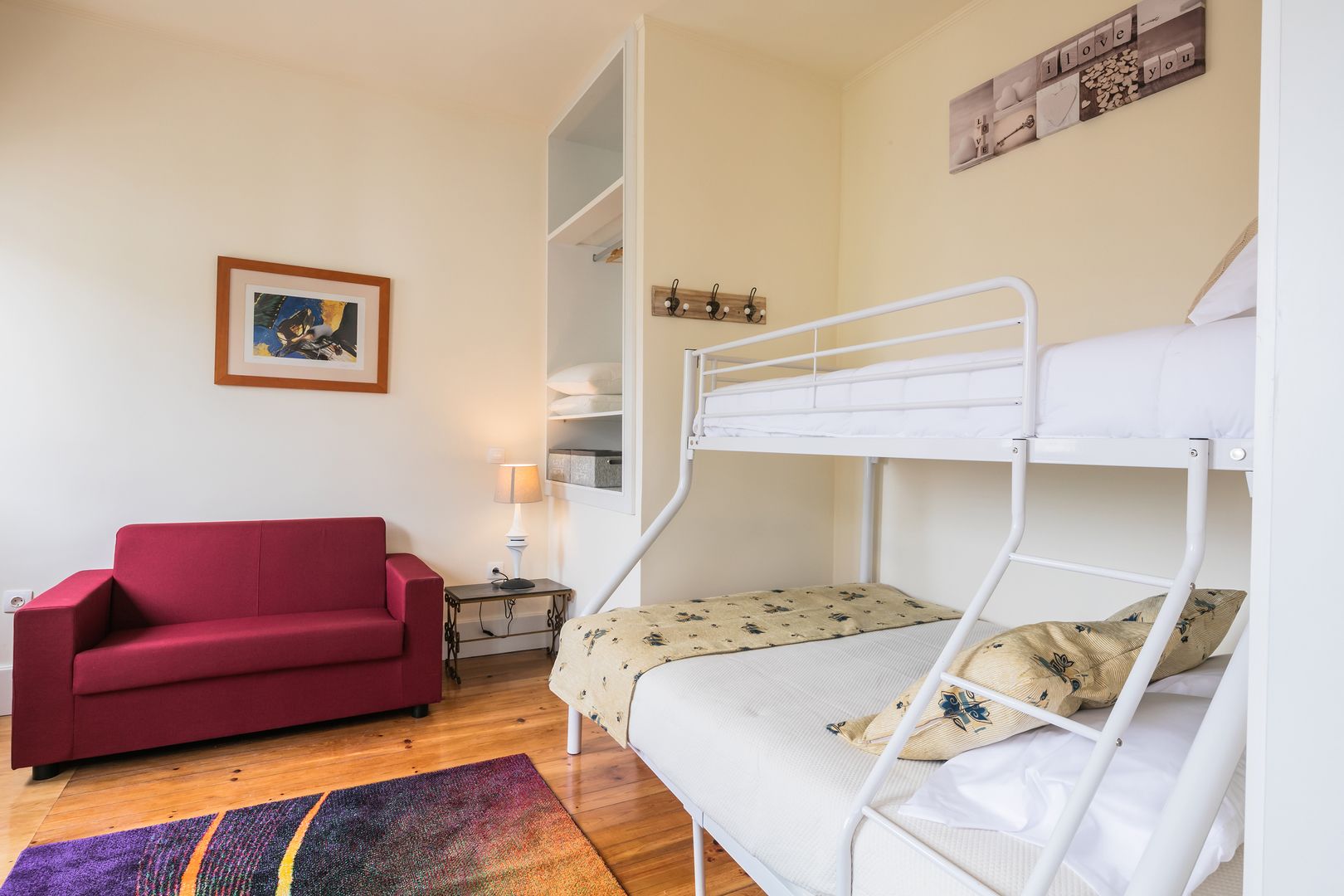 Una habitación con una cama litera, un sofá rojo, una mesita con una lámpara, un cuadro en la pared y una alfombra colorida.