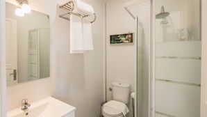 Ein Badezimmer mit Waschbecken, Toilette und Dusche. Ein Handtuchhalter mit einem Handtuch und ein kleines Regal mit Toilettenartikeln sind vorhanden.