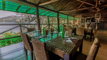 An outdoor dining area with wicker chairs and tables, overlooking a body of water and hills.
