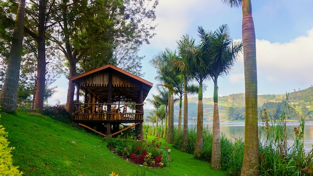 A wooden gazebo with a sloped roof, surrounded by lush greenery and palm trees, overlooking a calm lake with hills in the background. - Bunyonyi Overland Resort (Kabale)