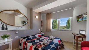 Ein Schlafzimmer mit einem großen Bett, einem Schreibtisch, einem Sessel, einem Fernseher und einem Fenster mit Blick auf Bäume.