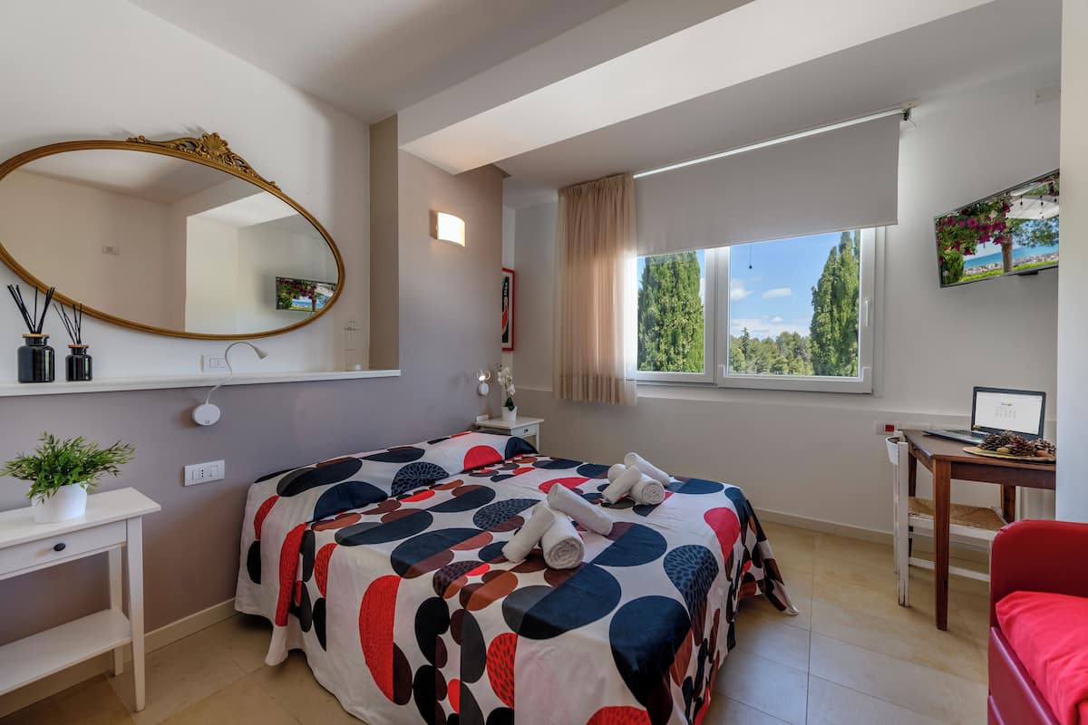 Een slaapkamer met een groot bed, een bureau, een stoel, een televisie en een raam met uitzicht op bomen.