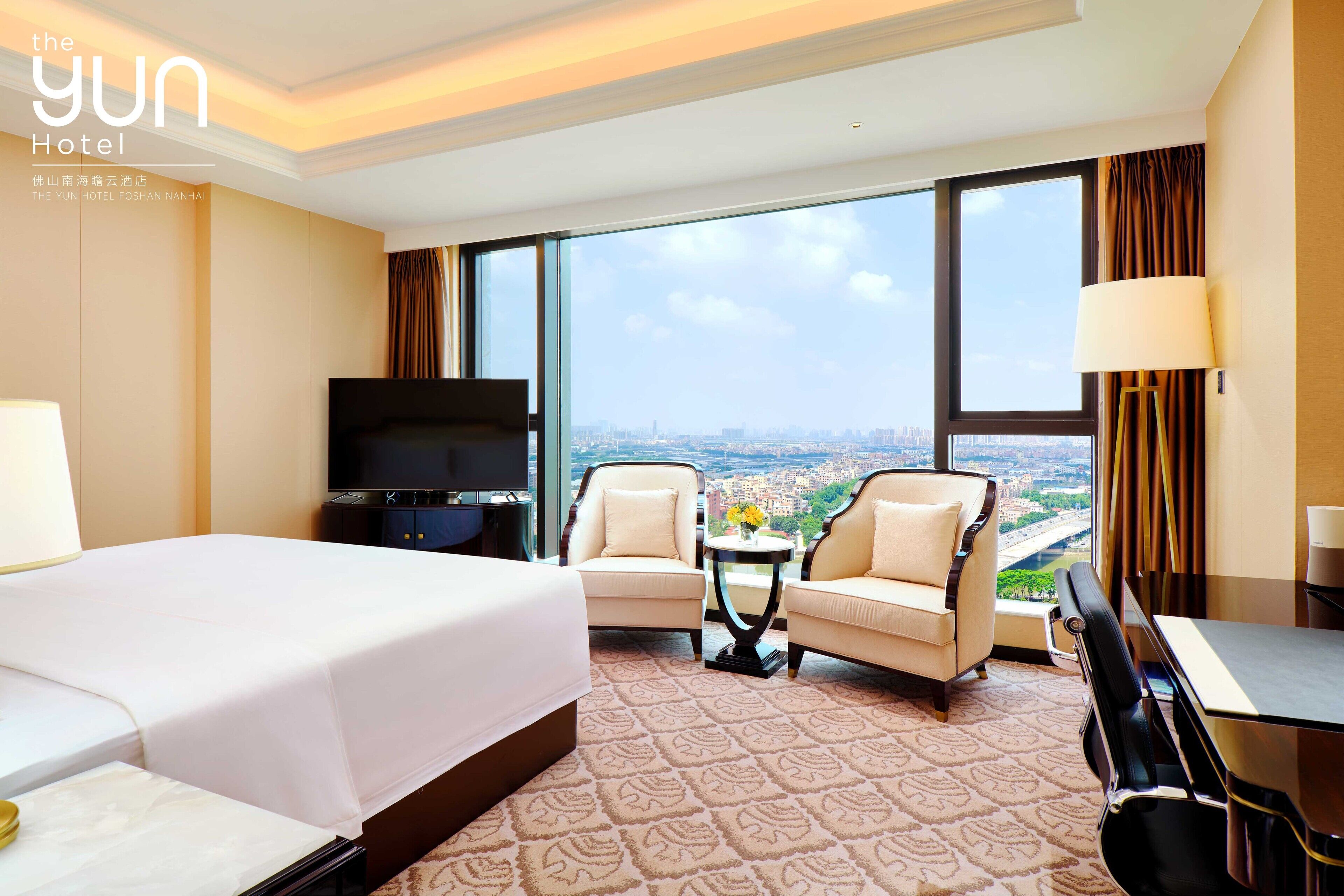 Habitación de hotel con una cama grande, dos sillones, una mesita, un televisor y vistas a la ciudad.