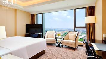 Habitación de hotel con una cama grande, dos sillones, una mesita, un televisor y vistas a la ciudad.