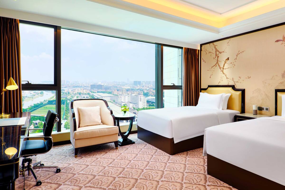 Une chambre d’hôtel avec deux lits, un bureau, une chaise et une grande fenêtre donnant sur la ville.