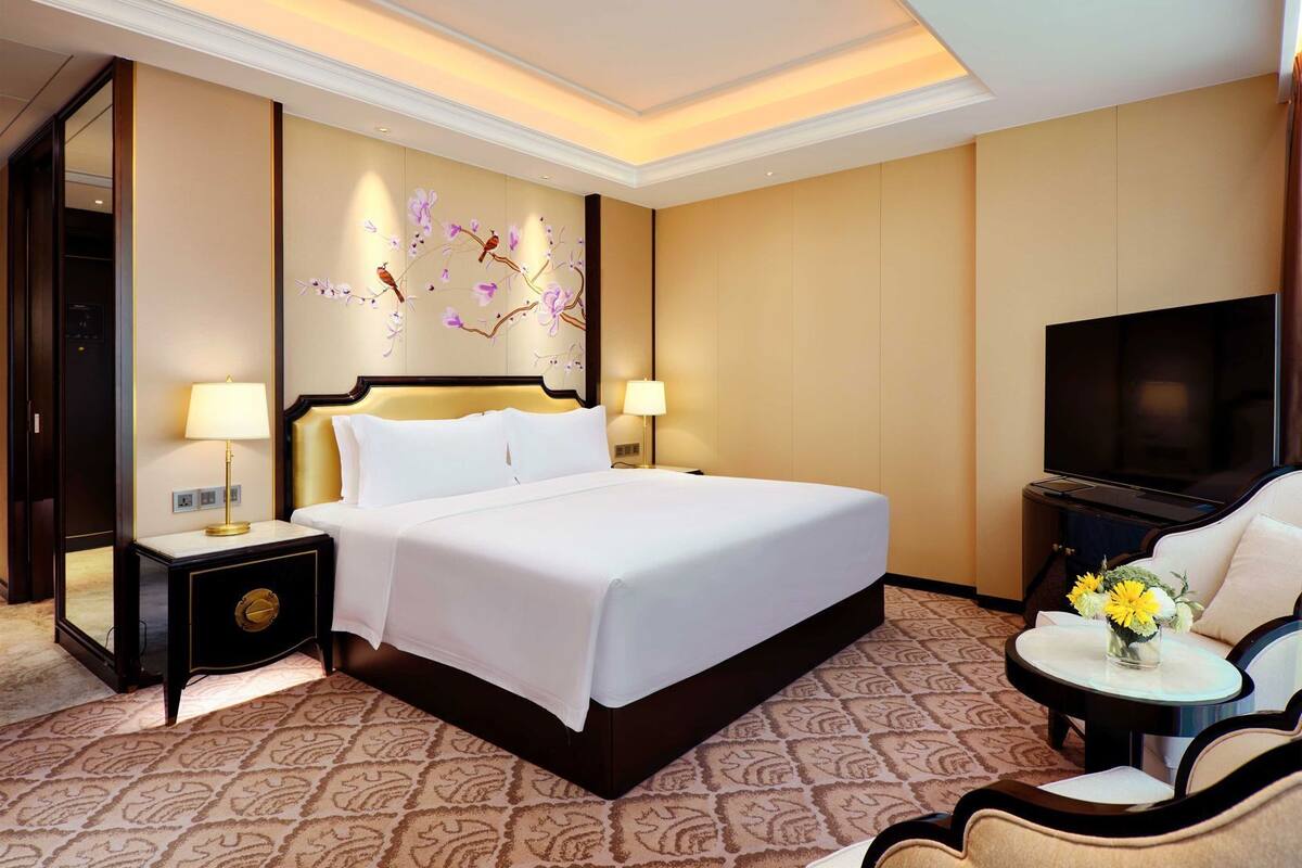 Une chambre d’hôtel avec un grand lit, une télévision, une chaise et une petite table agrémentée de fleurs.