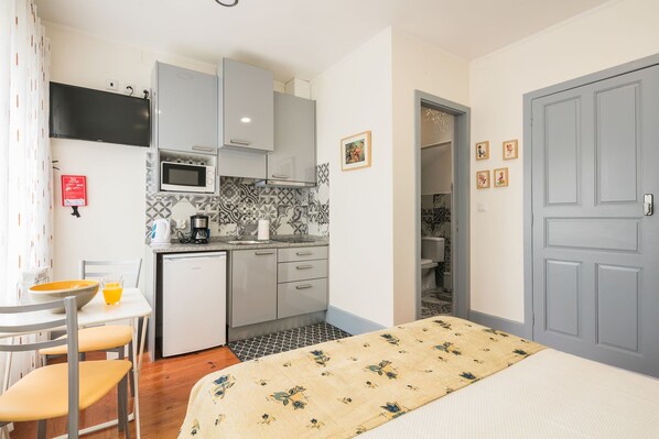 A compact hotel room with a kitchenette, a dining area with a table and chairs, a bed with a patterned bedspread, and a gray door. - Apartamento A Francos Burguer Station 2 (Porto)