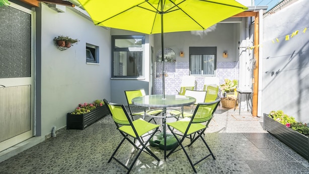 Un patio con una mesa redonda de vidrio y cuatro sillas verdes, un paraguas amarillo y una pared de azulejos con un diseño azul.