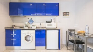 Cocina compacta con armarios azules, lavadora, lavavajillas, microondas y un pequeño comedor con una silla.