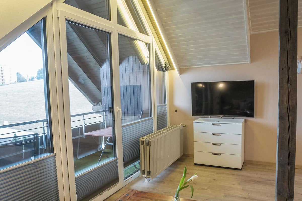 A room with a large sliding glass door leading to a balcony, a wooden table with a bowl and glass, a white dresser, and a flat-screen TV mounted on the wall.