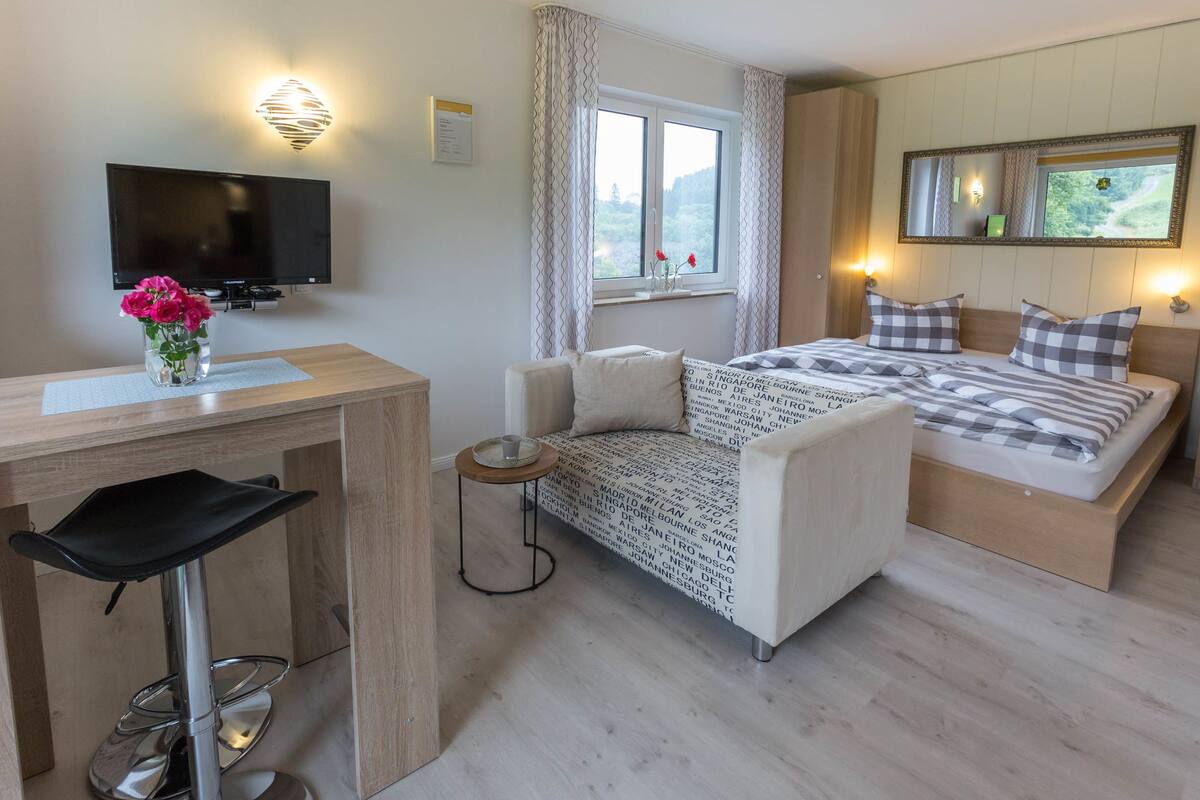 A modern hotel room with a bed, a desk with a chair, a TV, and a window with curtains.