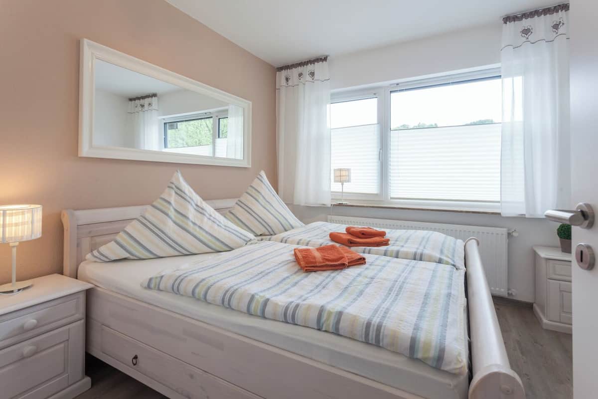 A bedroom with two beds, a nightstand, a dresser, a window with curtains, and a radiator.
