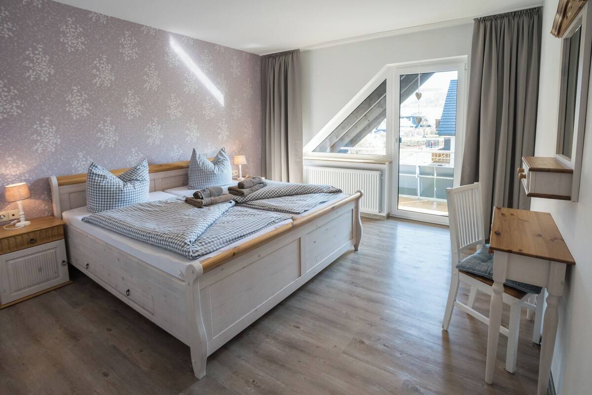 A bedroom with a large bed, a desk, and a chair. There is a window with curtains and a view of buildings outside.