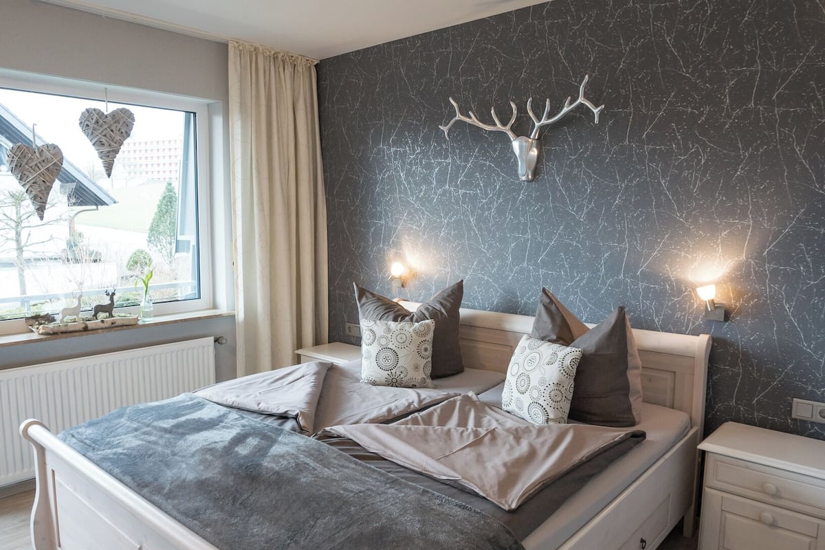 A bedroom with a bed, bedside tables, a window with curtains, a wall-mounted deer head, and a view of the outside.