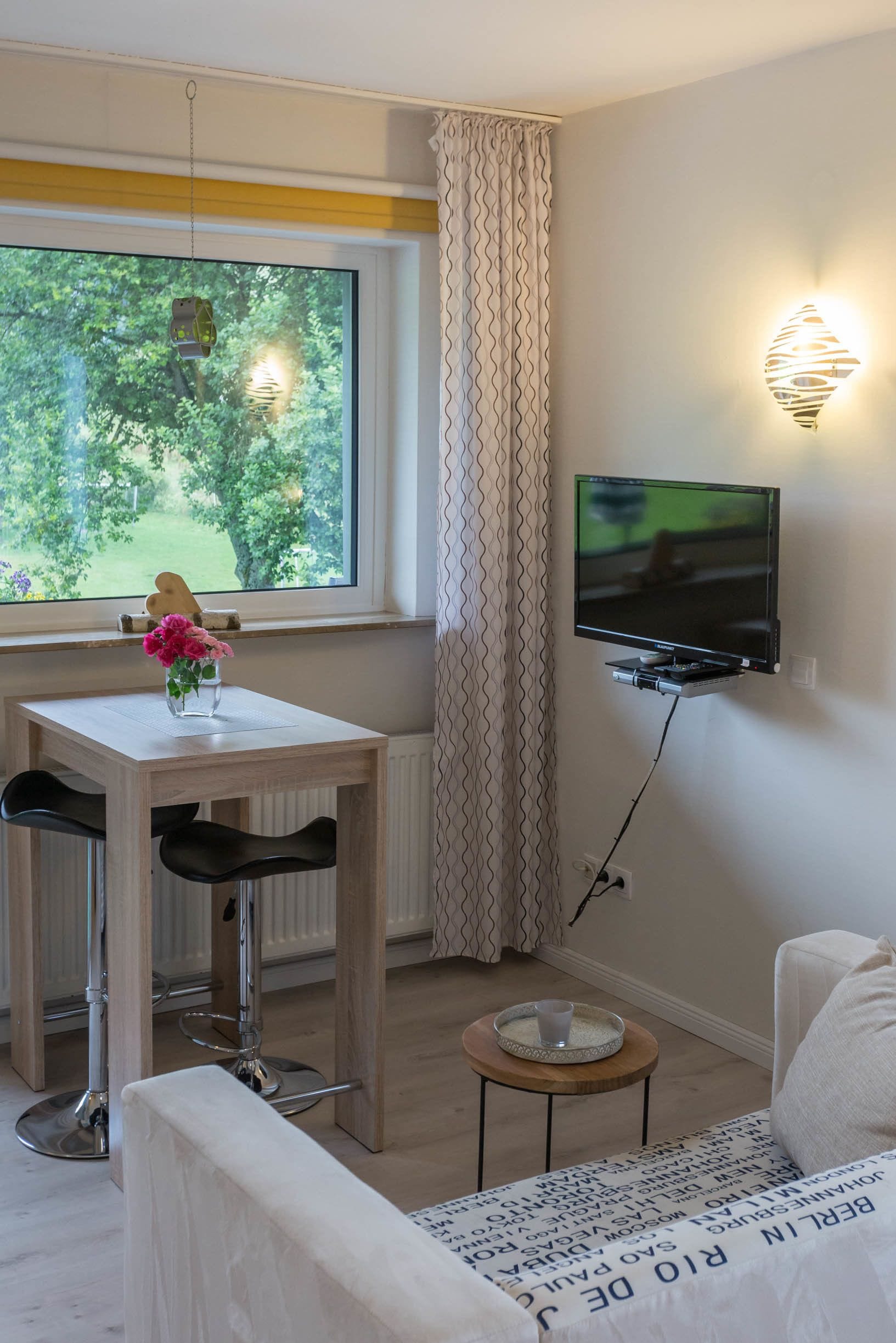 A room with a wooden table, a TV mounted on the wall, a window with curtains, and a small table with a cup.