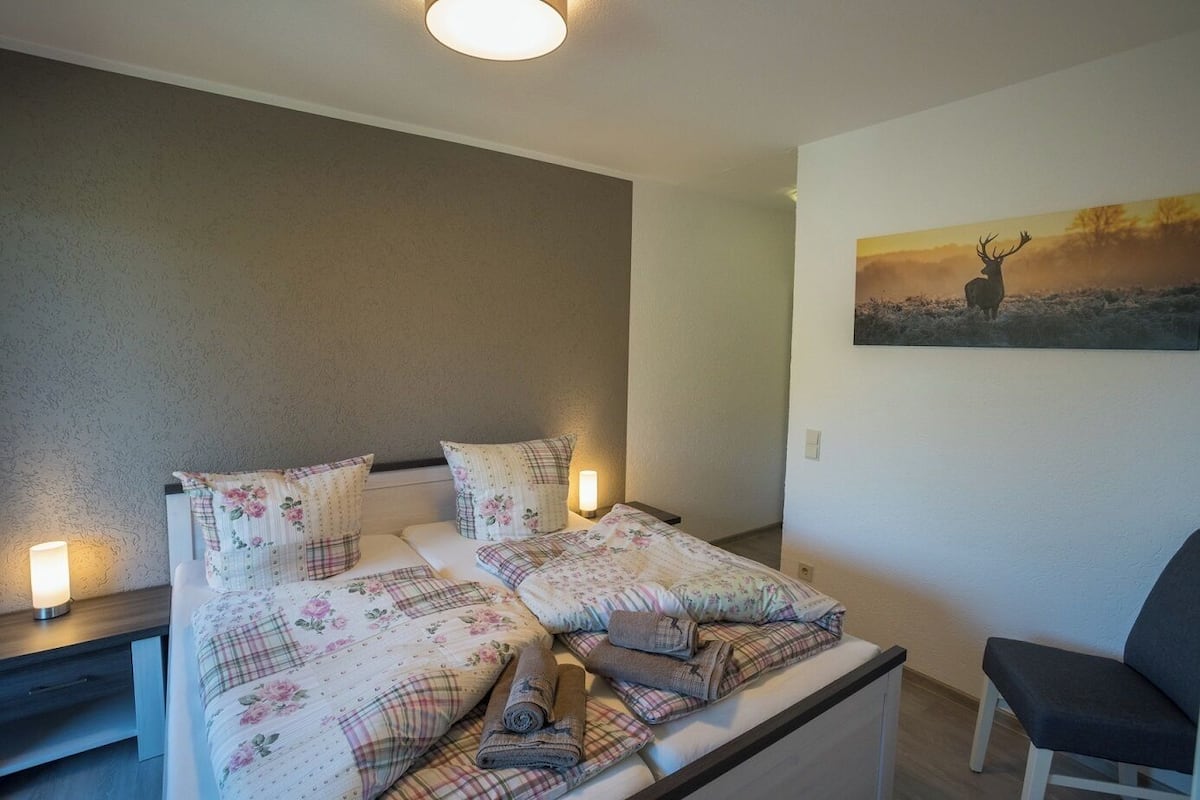 A bedroom with two beds, a nightstand, a chair, and a painting of a deer on the wall.