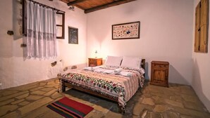 A bedroom with a stone floor, a bed with a floral bedspread, a nightstand, a wooden cabinet, and a wall-mounted picture. - Stone made Mansions (Apokoronas)