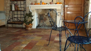 A room with a fireplace, a metal chair, a metal table, a metal shelf with books and a plant, and a wooden door. - Chambres d'hôtes Côté Jardin (Villiers-sur-Loir)