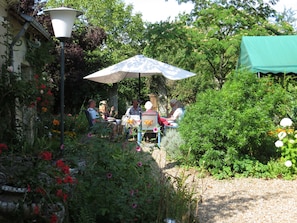 Un patio jardín con una mesa y sillas bajo un paraguas grande, rodeado de vegetación exuberante y plantas con flores.