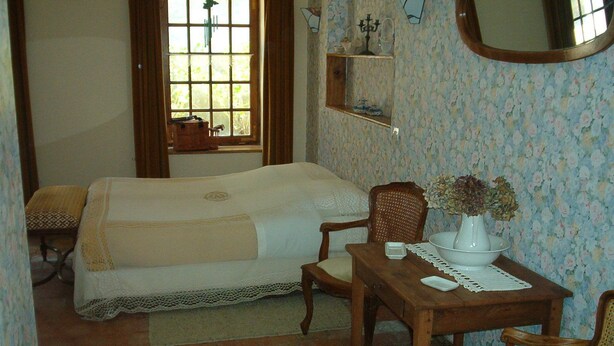 Un dormitorio con cama, dos sillas, una mesita con un jarrón, una ventana y una estantería decorada.