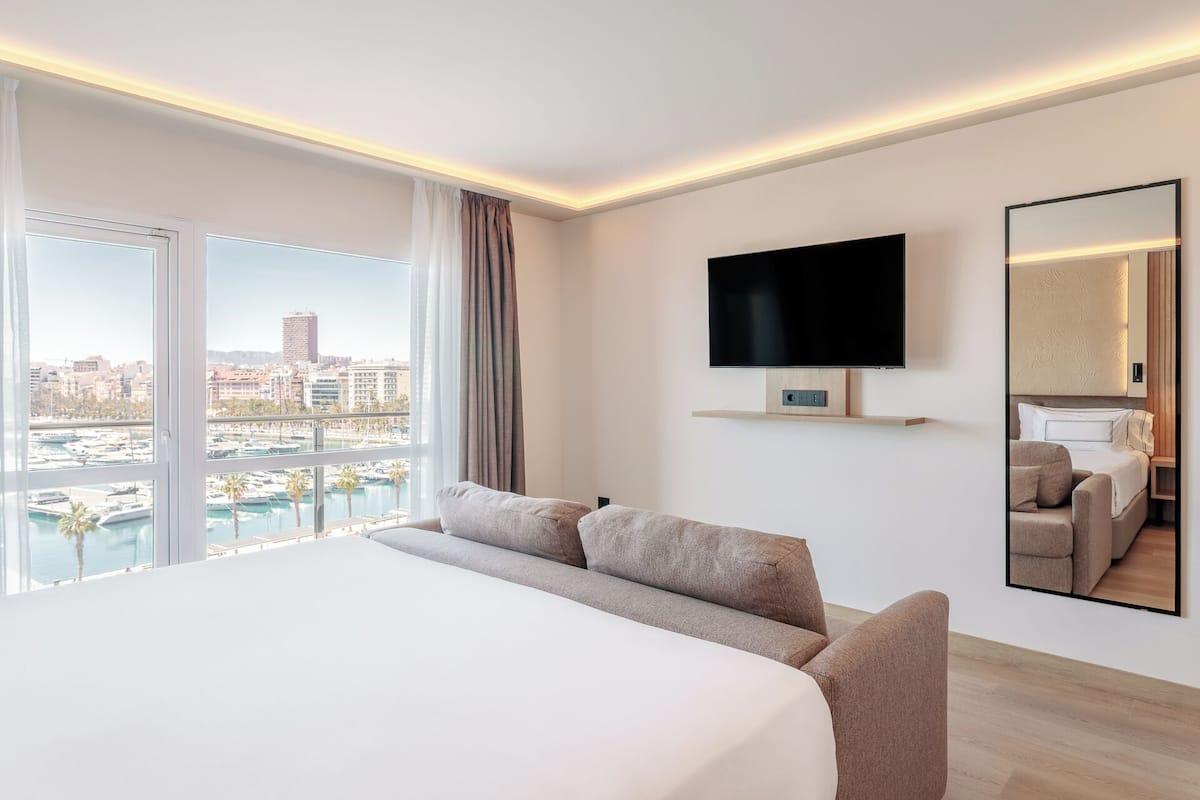 Een moderne hotelkamer met een groot bed, een aan de muur bevestigde televisie, een spiegel en uitzicht op de stad en jachthaven door grote ramen.
