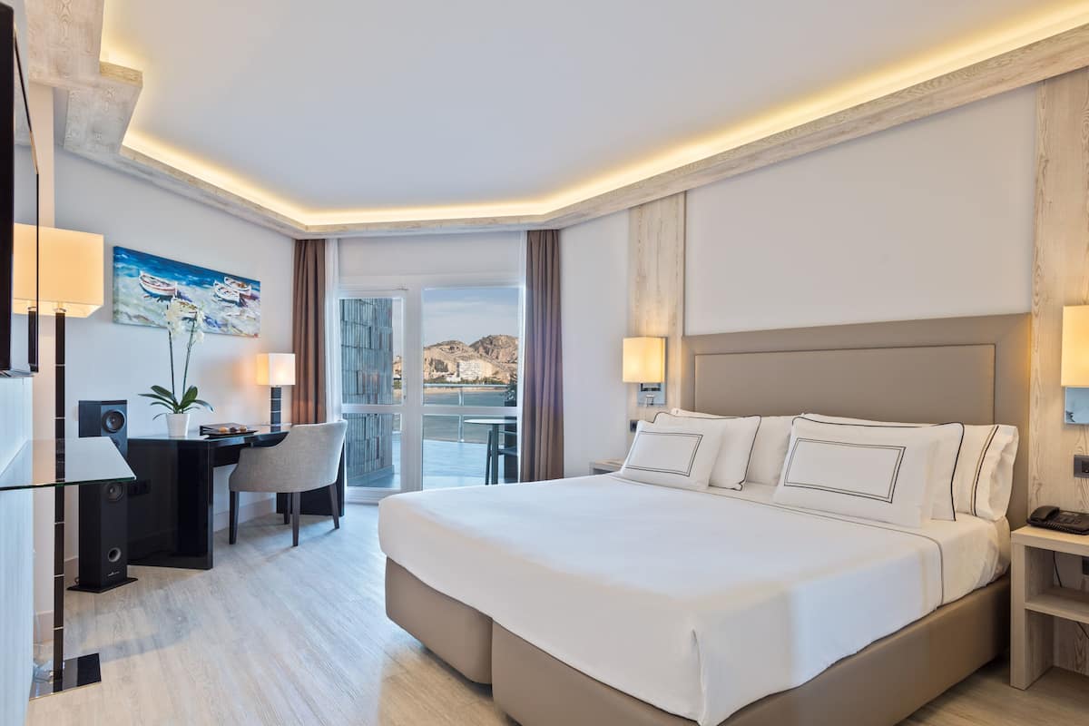 Een hotelkamer met een groot bed, een bureau met stoel, een bureaulamp, een schilderij aan de muur en uitzicht op het omliggende landschap.