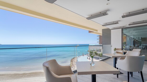 A modern dining area with a view of the ocean, featuring a table and chairs, a vase with flowers, and a glass railing overlooking the beach. - The Level at Melia Alicante - Adults Only (Alicante)