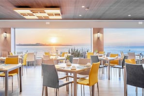 A restaurant with tables and chairs arranged for dining, large windows offering a view of the sunset over the water.