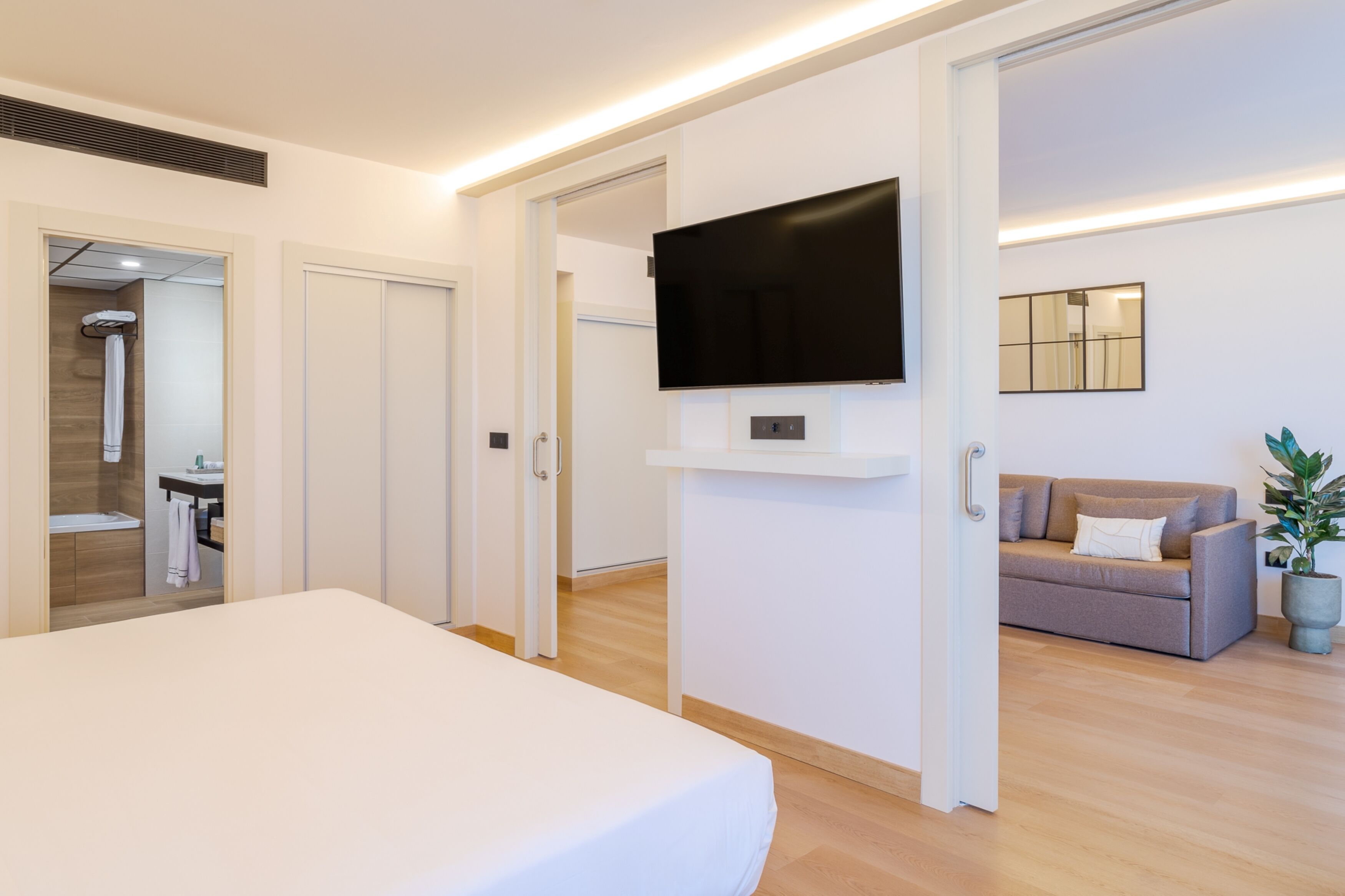 Ein modernes Hotelzimmer mit einem großen Bett, einem flachbildfernseher an der Wand, einer Couch und einer Zimmerpflanze.
