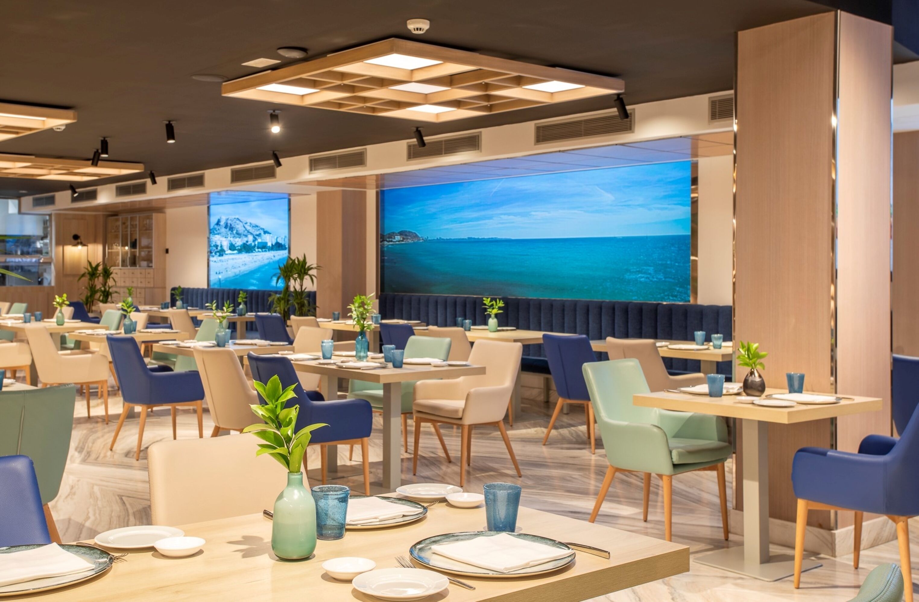 Un restaurante moderno con varias mesas y sillas, amplias ventanas con vista al mar e iluminación empotrada en el techo.