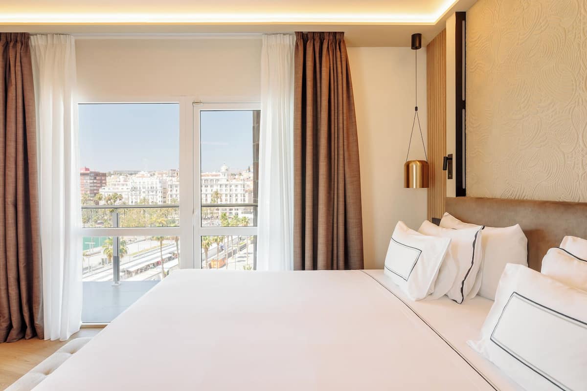 Hotelkamer met een groot bed, uitzicht op de stad door middel van kamerhoge ramen en een modern design met een gestructureerde wand en een hanglamp.