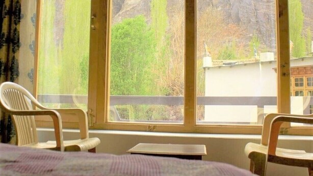 Ein Zimmer mit großem Fenster und Blick auf Berge und grüne Landschaft. Ein Bett mit Decke und zwei Stühle sind vorhanden.