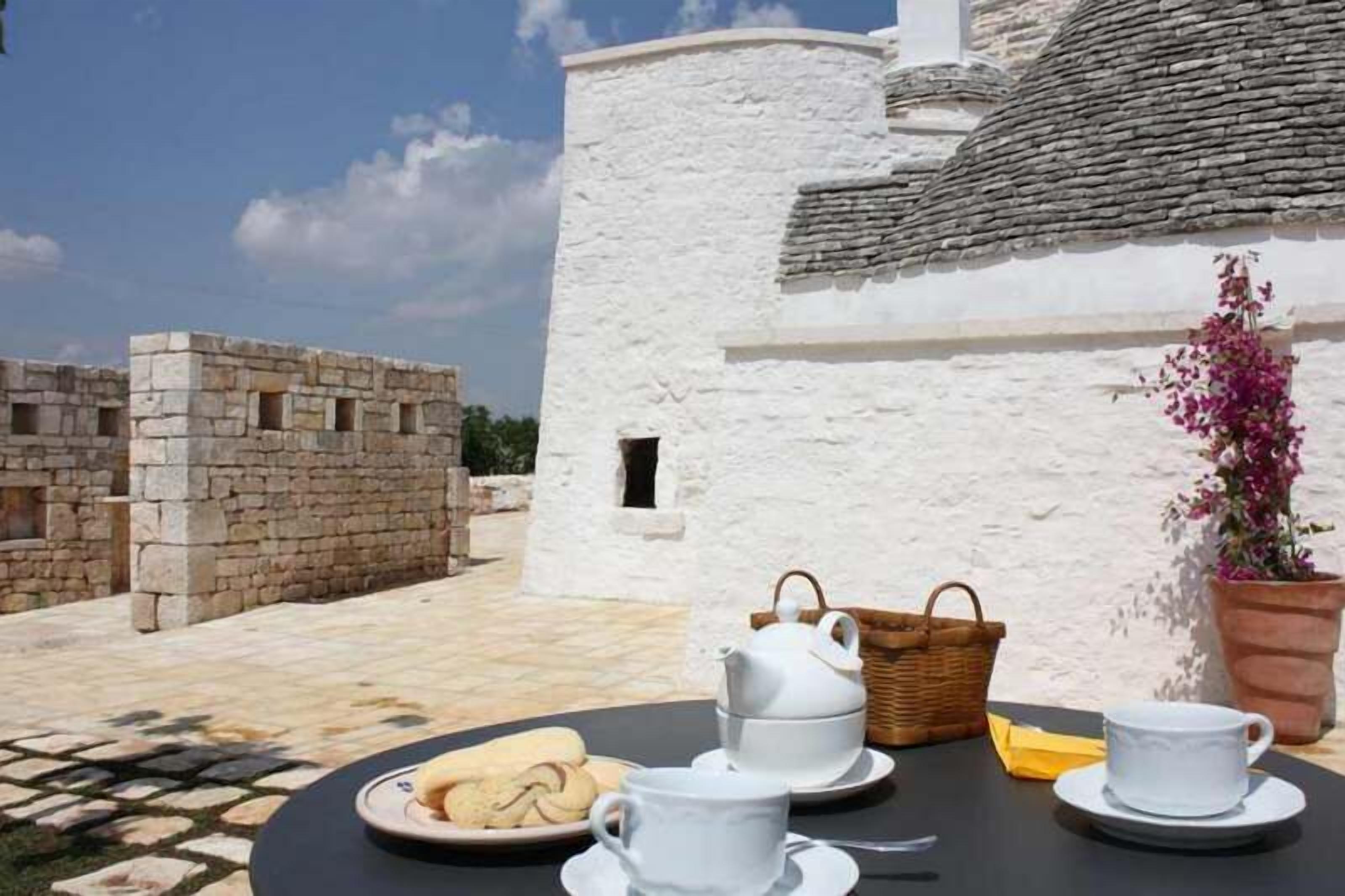 Une table dressée avec une théière, des tasses et un panier, avec un bâtiment blanc et un mur de pierre en arrière-plan.