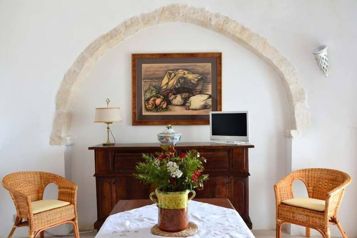 Um quarto com uma mesa de jantar, duas cadeiras de vime, um armário de madeira, uma televisão, um quadro e um vaso com flores.