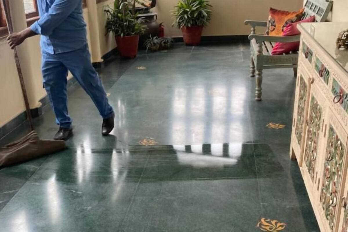 A person walking in a hallway with a brown mat on the floor, potted plants, and a wooden bench with cushions.