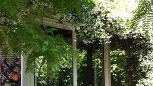 A glass-enclosed outdoor seating area with a table and chairs, surrounded by lush greenery and trees.