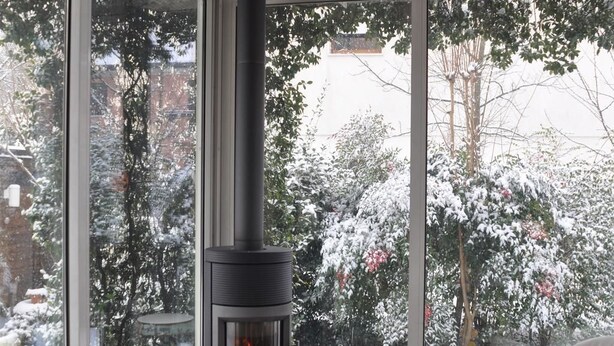 A room with a large window overlooking a snowy garden, a fireplace, and a table with a kettle.