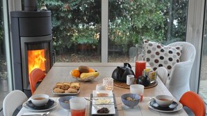 A cozy breakfast setup with a wood-burning stove, a table set with fruits, bread, and beverages, and a view of greenery through large windows.