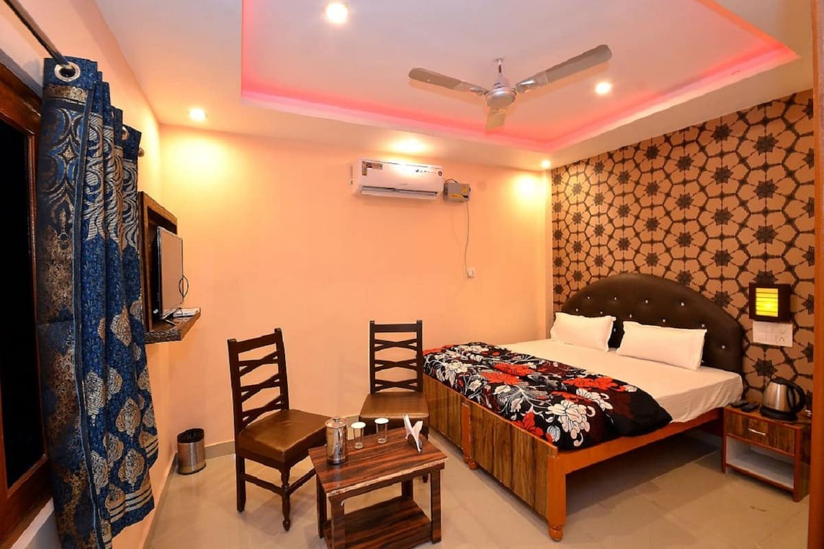 A bedroom with a bed, a desk, chairs, a ceiling fan, an air conditioner, and a nightstand with a lamp.
