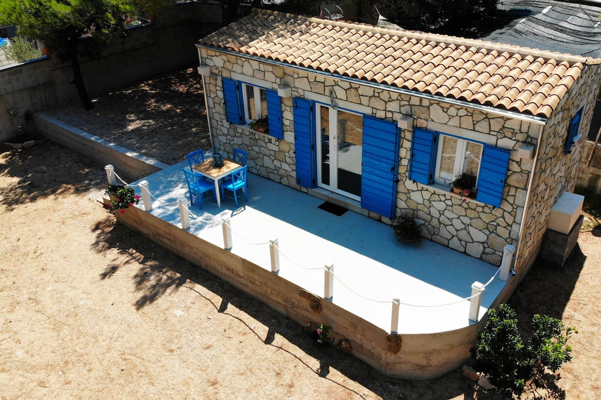 A stone house with a tiled roof, a small patio with a table and chairs, and a fenced area.