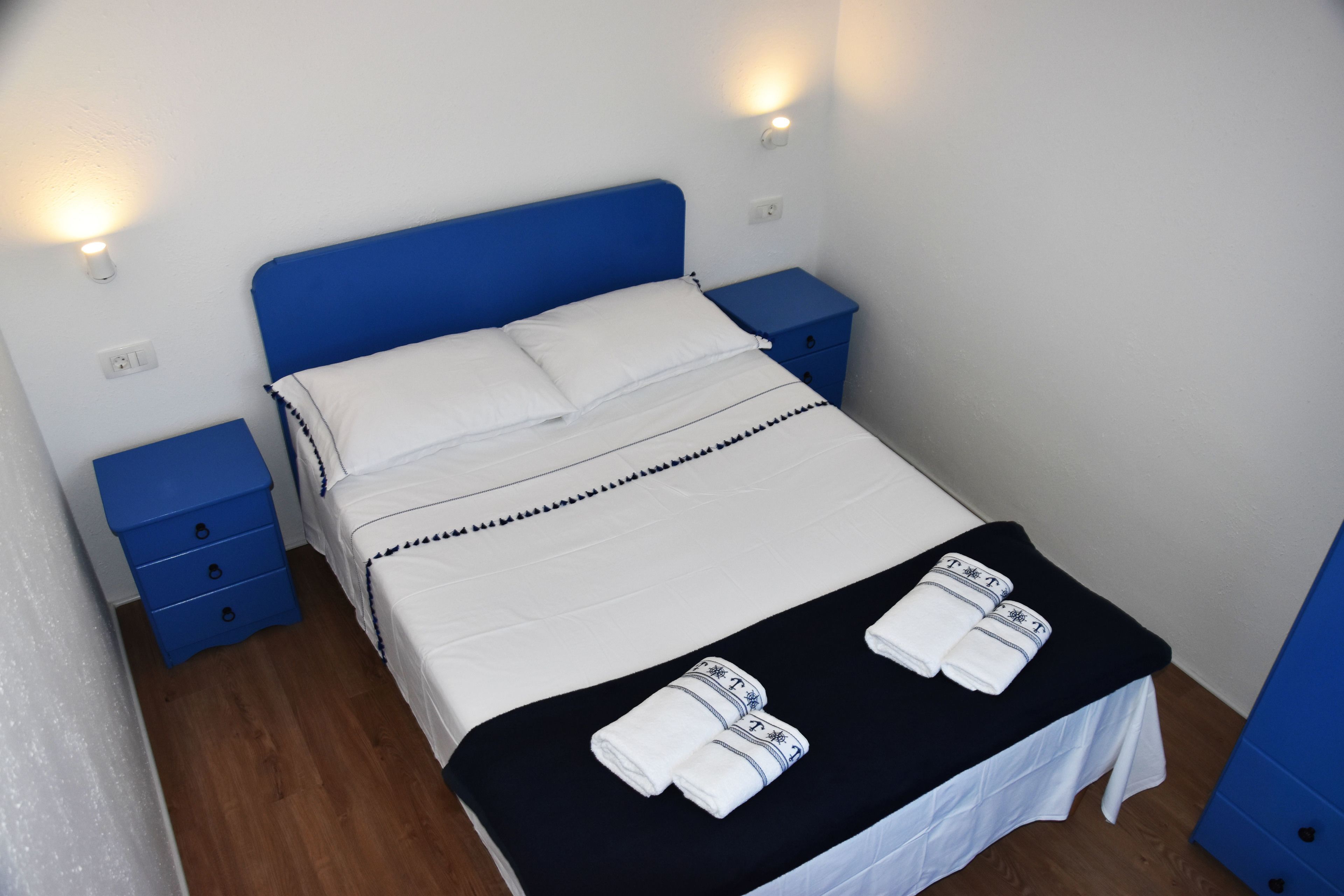 A neatly made bed with white linens and a black bedspread, two blue nightstands with drawers, and a wall-mounted light fixture.