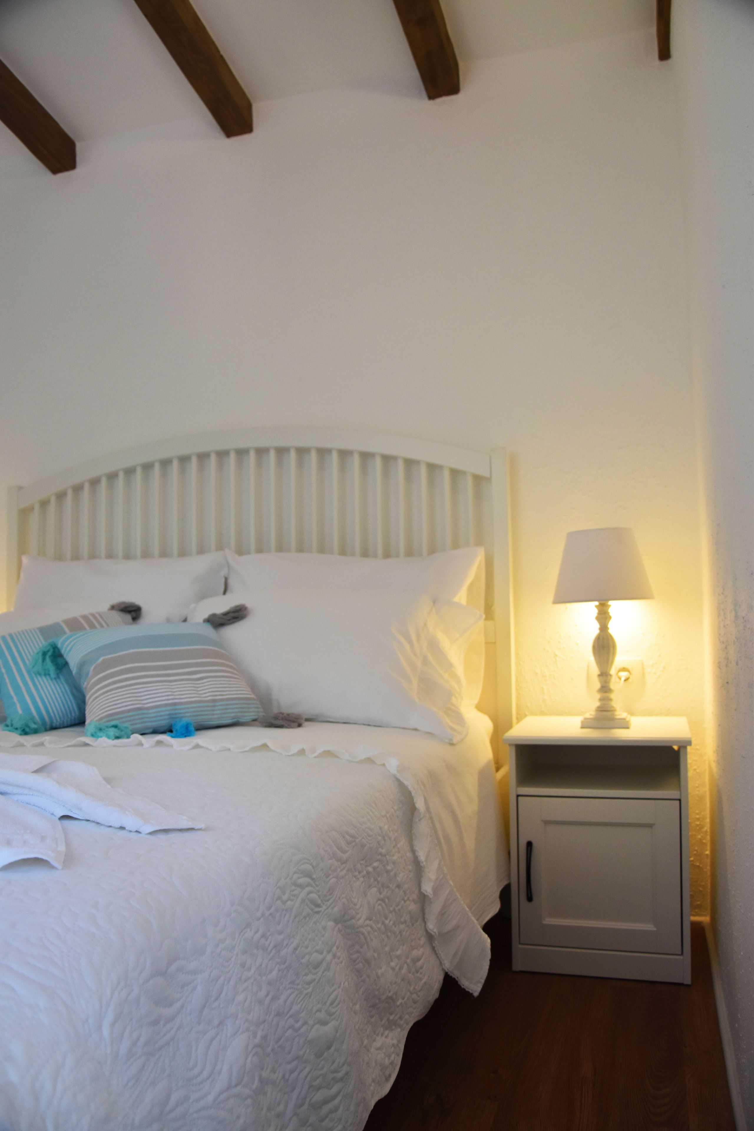 Una cama bien hecha con ropa de cama blanca y varios almohadones decorativos, una mesita de noche con lámpara y suelo de madera.