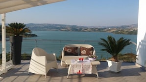 A terrace with a sofa and chairs, a table with glasses and a bowl, and a view of a lake and hills.